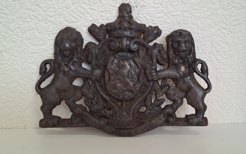 May include: A cast iron decorative plaque featuring two lions flanking a shield with a crown above. The plaque is in a dark brown color and has a distressed finish.