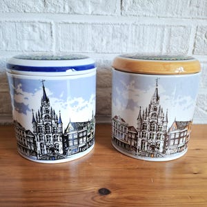 Può includere: Due barattoli in ceramica con coperchi. Ogni barattolo presenta una dettagliata illustrazione in bianco e nero di un paesaggio urbano europeo su uno sfondo di cielo azzurro. I coperchi hanno un bordo decorativo.