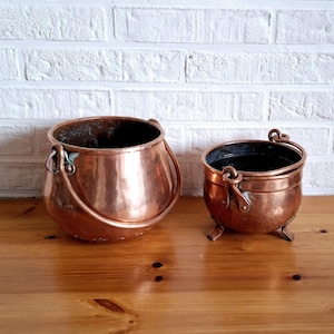 Peut inclure: Deux pots en cuivre, l'un plus grand que l'autre, avec des poignées. Le plus grand pot a un fond arrondi et le plus petit pot a trois pieds.