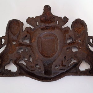 May include: A cast iron wall plaque with a coat of arms design. The plaque features two lions flanking a shield with a crown above. The plaque is rusty brown in color.