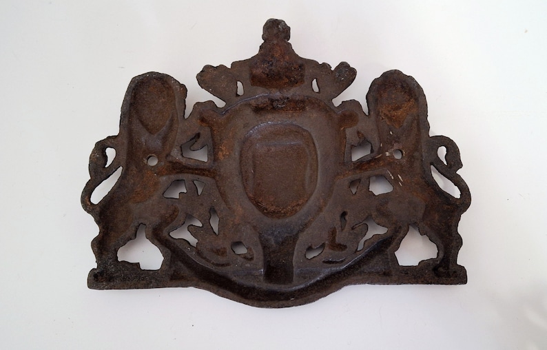 May include: A cast iron wall plaque with a coat of arms design. The plaque features two lions flanking a shield with a crown above. The plaque is rusty brown in color.