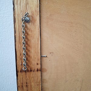 May include: Close-up of a wooden frame with a metal chain and screw attached. The frame is made of light brown wood, and the chain is silver. A small nail is also visible on the frame.