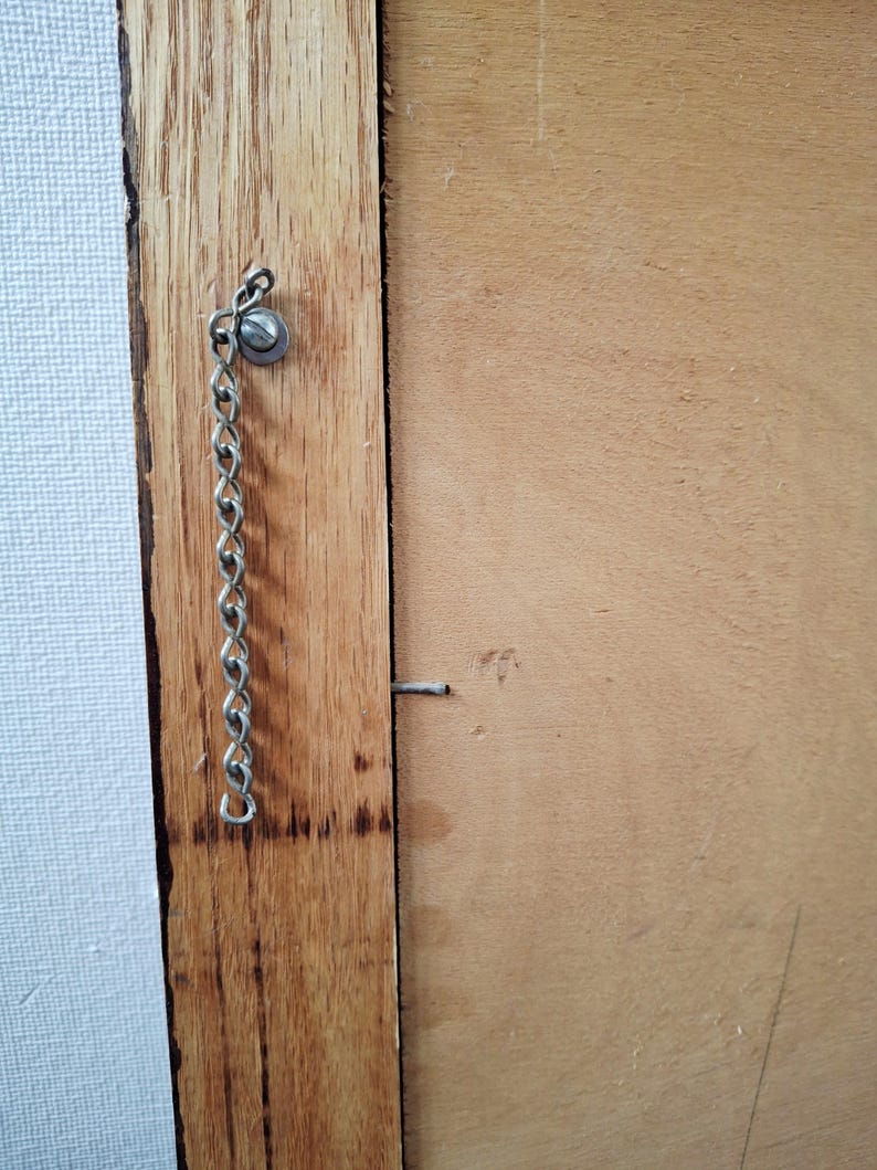 May include: Close-up of a wooden frame with a metal chain and screw attached. The frame is made of light brown wood, and the chain is silver. A small nail is also visible on the frame.