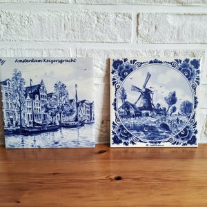 Può includere: Due piastrelle in ceramica blu e bianca. Una piastrella raffigura una scena di canale con edifici e barche, con il testo "Amsterdam Keizersgracht". L'altra piastrella mostra un mulino a vento, un ponte e case. Entrambe hanno un bordo floreale.