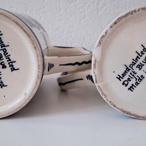 May include: Two white ceramic mugs with blue and black hand-painted designs. The bottom of each mug reads "Handpainted Delft Blue Made in Holland."