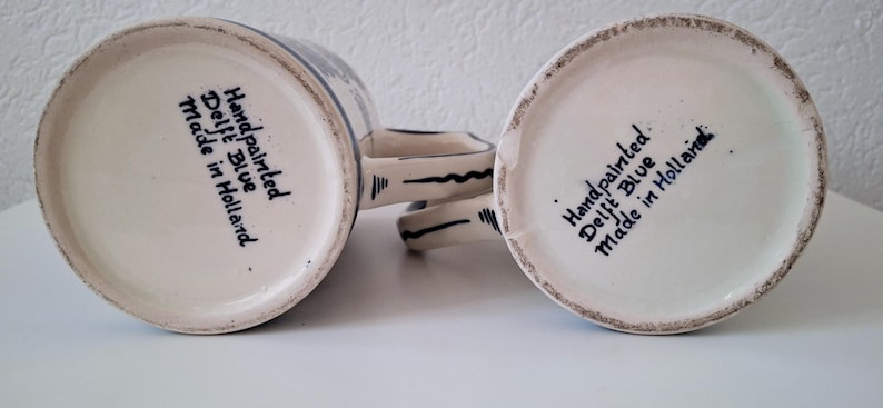 May include: Two white ceramic mugs with blue and black hand-painted designs. The bottom of each mug reads "Handpainted Delft Blue Made in Holland."