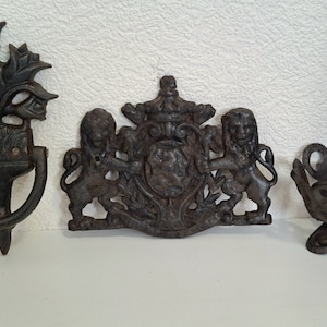 May include: Three antique cast iron wall decor pieces. The first is a door knocker with a floral design. The second is a coat of arms with two lions. The third is a small decorative bowl.