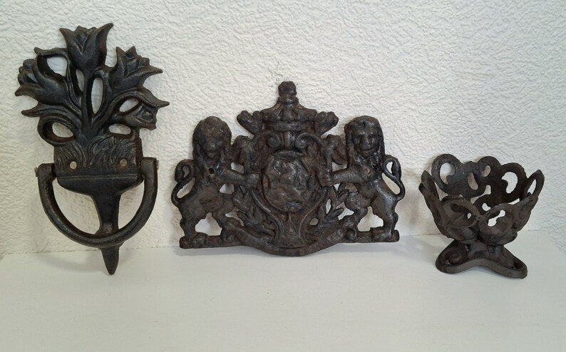 May include: Three antique cast iron wall decor pieces. The first is a door knocker with a floral design. The second is a coat of arms with two lions. The third is a small decorative bowl.