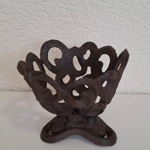 May include: A dark brown cast iron candle holder with a decorative, intricate design. The holder has a wide, open top and a three-legged base.