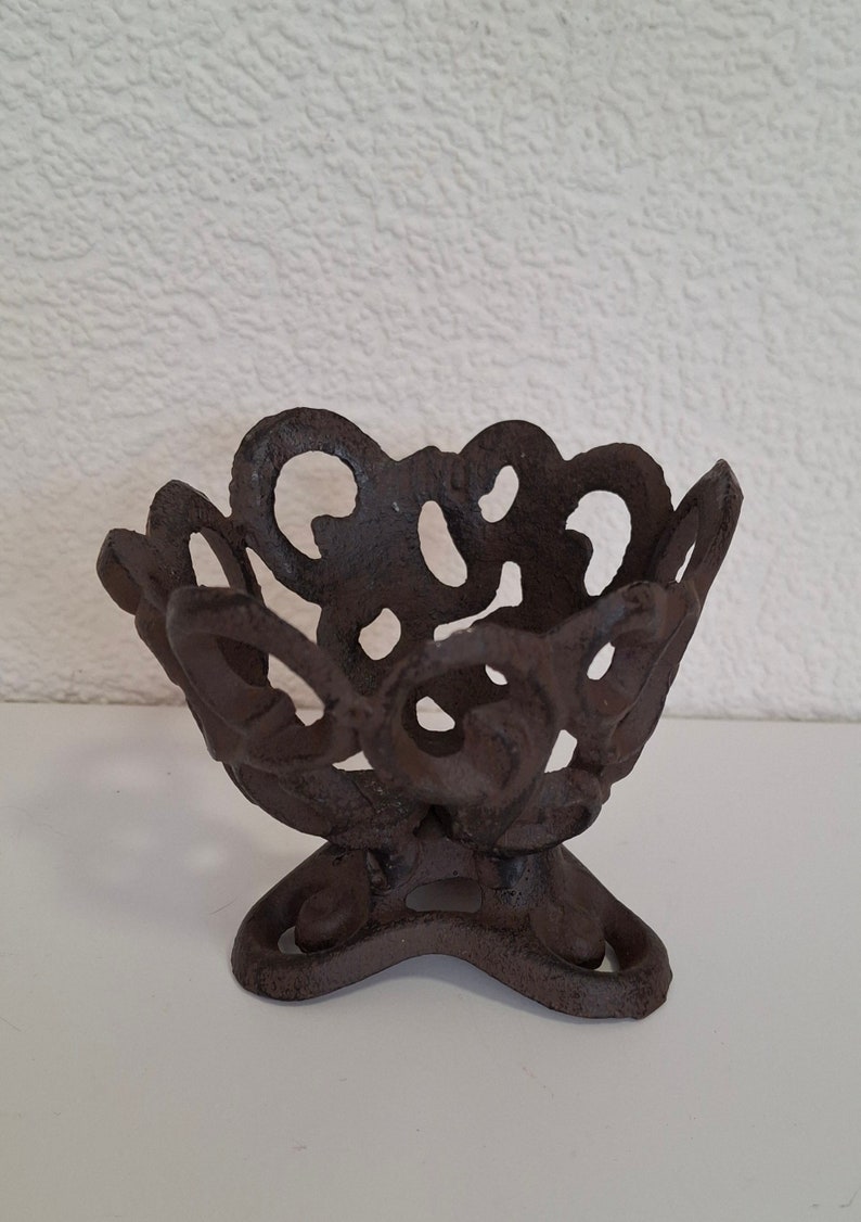 May include: A dark brown cast iron candle holder with a decorative, intricate design. The holder has a wide, open top and a three-legged base.