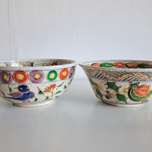 Op de afbeelding: Twee decoratieve keramische kommen met handgeschilderde bloemen- en vogelontwerpen. De kommen hebben een craquelé glazuur en zijn versierd met kleurrijke patronen van bloemen, cirkels en vogels. De kommen staan op een wit oppervlak.