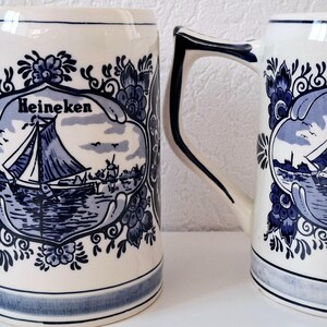 May include: Two white ceramic beer mugs with blue and white Delftware designs. The mugs feature a sailboat with the word "Heineken" on each.