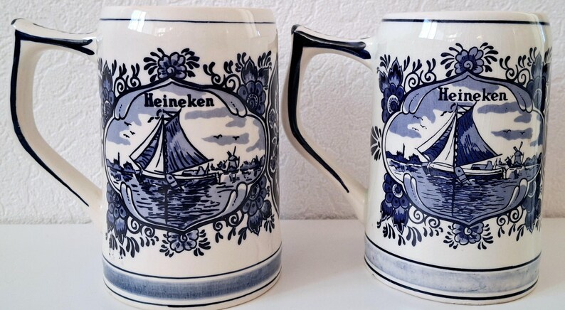 May include: Two white ceramic beer mugs with blue and white Delftware designs. The mugs feature a sailboat with the word "Heineken" on each.