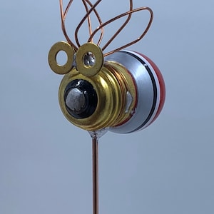May include: A handcrafted bee sculpture made from repurposed materials. The bee features a light bulb body with red, white, and black stripes, brass washers for eyes, and copper wire wings. It is mounted on a copper rod.
