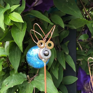 May include: A whimsical garden art piece featuring a blue glass orb body, copper wire wings, and brass hardware details. The decorative insect sculpture is mounted on a copper rod, set against a backdrop of green leaves and purple flowers.