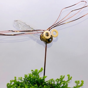 May include: A dragonfly sculpture made from copper wire and a light bulb. The dragonfly has a clear glass body and gold-colored accents. The sculpture is perched on a thin metal rod.