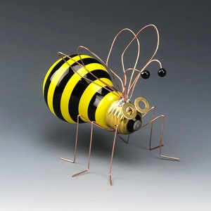 May include: A yellow and black striped light bulb bee with copper wire legs and antennae. The bee has two small black eyes and a copper wire mouth.