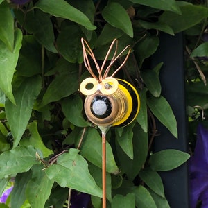 May include: A whimsical garden decoration featuring a bee made from recycled materials. The bee has a yellow and black striped body, copper wire antennae, and large, round, black eyes with yellow rims.
