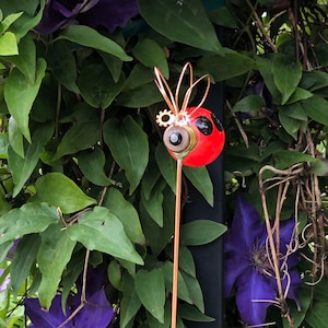 May include: A red and black ladybug garden stake with copper wire wings and gears. The stake is surrounded by purple flowers and green leaves.