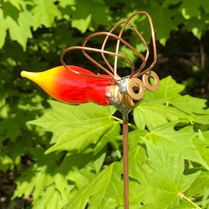 May include: A red and yellow glass hummingbird with copper wire wings and gold metal eyes. The hummingbird is attached to a copper colored metal stick.