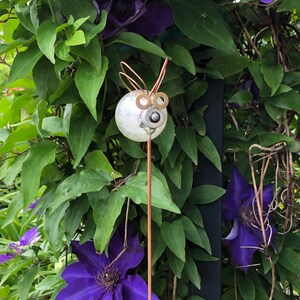 May include: A white metal garden stake with a whimsical face and copper wire ears. The stake is surrounded by purple clematis flowers and green leaves.