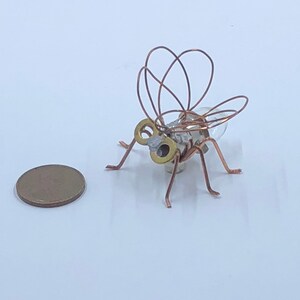 May include: A small, copper wire sculpture of a fly with a clear glass body and gold accents. The sculpture is sitting on a white surface.