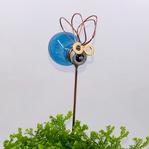 May include: A blue glass lightbulb with copper wire antennae and gold metal eyes, mounted on a metal rod and placed in a pot of green foliage.
