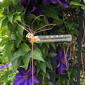 May include: A copper wire dragonfly sculpture with a light bulb body, mounted on a metal rod. The dragonfly is set against a backdrop of green leaves and vibrant purple flowers.