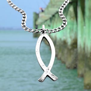 Puede incluir: Un collar de cadena de plata con un colgante de pescado de plata. El colgante es un diseño simple y abierto con una superficie texturizada.