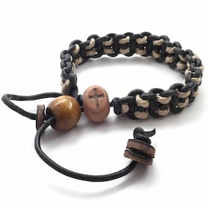 Op de afbeelding: Een zwart en wit gevlochten leren armband met een houten kraal en een kraal in de vorm van een kruis. De armband heeft een leren sluiting.