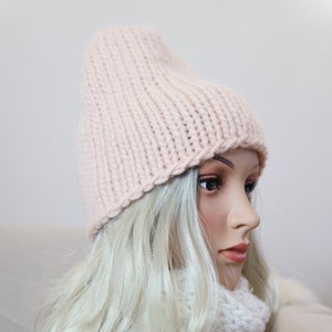 May include: A soft, pale pink knitted beanie hat with a folded brim. The hat is worn on a mannequin with long, blonde hair.