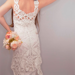 May include: A white crochet dress with a low back and a scalloped hem. The dress is worn by a person holding a bouquet of pink flowers.