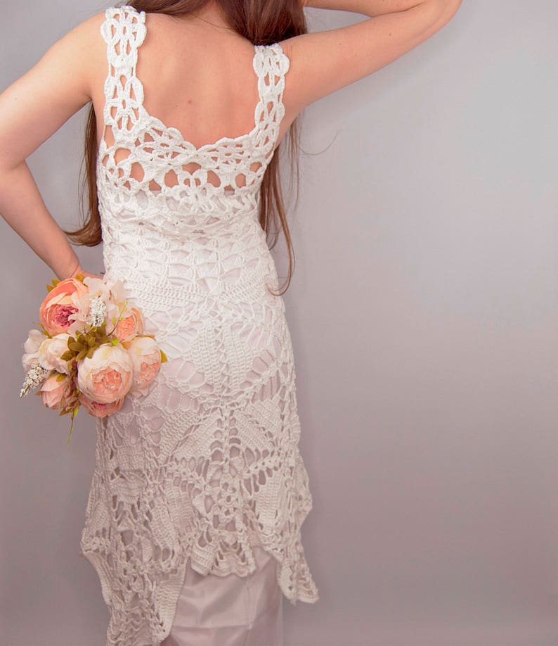 May include: A white crochet dress with a low back and a scalloped hem. The dress is worn by a person holding a bouquet of pink flowers.