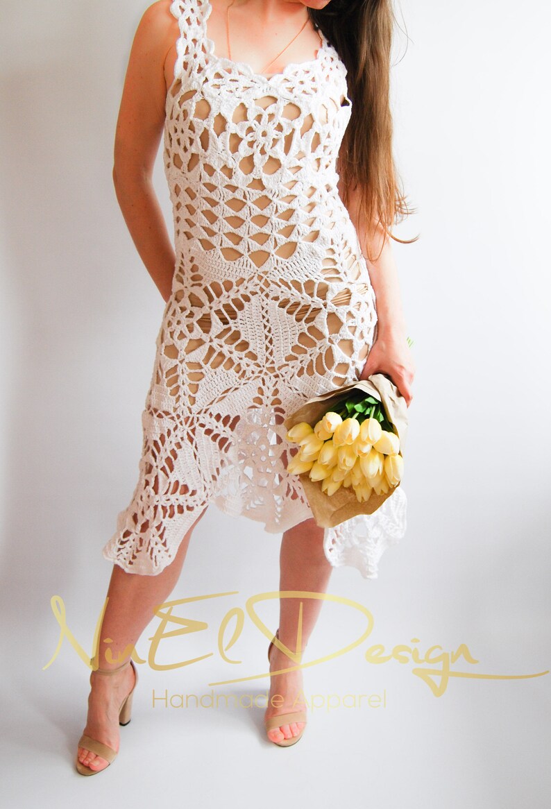 May include: White crochet dress with a delicate floral pattern. The dress has a loose fit and a flowing skirt. The model is wearing the dress with a bouquet of yellow flowers.