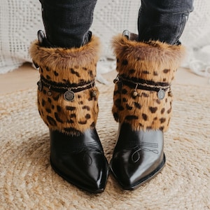 以下が含まれることがあります： ヒョウ柄のファーカフスと装飾的な金属製のアクセントが付いた、黒い革製のカウボーイブーツのペアです。