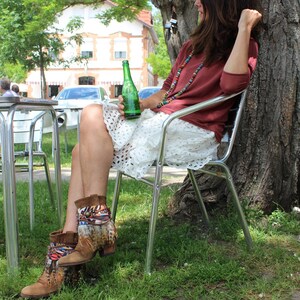 May include: A woman wearing a white lace skirt and a maroon top sits on a metal chair with a green glass bottle in her hand. She is wearing brown boots with colourful feathers and beads around her neck.