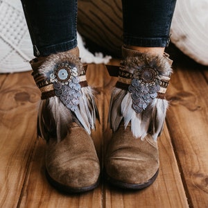 Könnte beinhalten: Braune Wildleder-Stiefeletten mit Feder- und Perlenbesatz. Die Stiefeletten haben ein dekoratives Band mit silbernen Münzen und einem braunen Lederriemen.