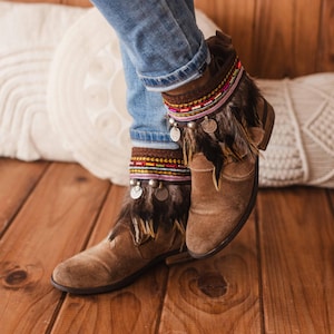 May include: Brown suede ankle boots with decorative feather and beaded trim. The boots are worn with blue jeans.