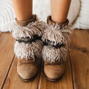 Op de afbeelding: Een paar enkellaarsjes van beige suède met een dikke, ruige rand van imitatiebont aan de bovenkant. Elke laars is voorzien van een donker leren bandje versierd met kleine, ronde metalen bedeltjes en studs voor extra textuur.