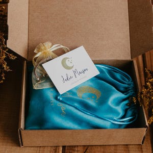 May include: A brown cardboard shipping box containing a bright turquoise satin drawstring pouch with a gold moon graphic, a small sheer yellow organza bag, and a white rectangular card featuring a gold moon logo and the text Indie Maison.