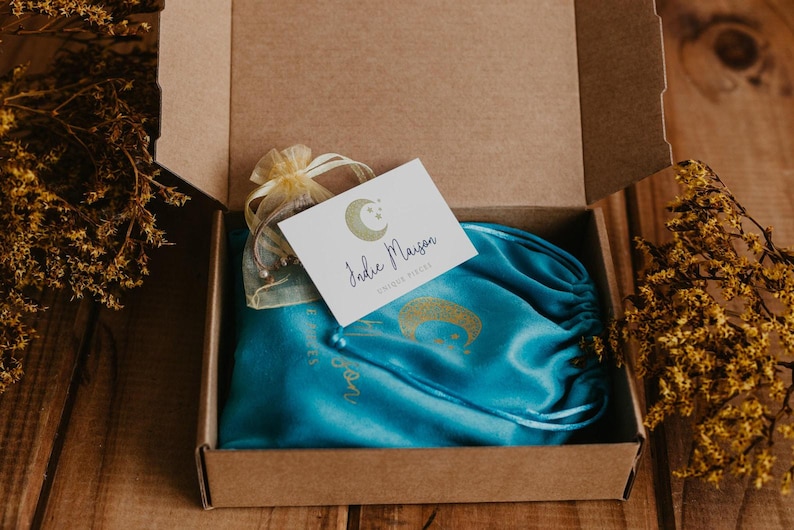 May include: A brown cardboard shipping box containing a bright turquoise satin drawstring pouch with a gold moon graphic, a small sheer yellow organza bag, and a white rectangular card featuring a gold moon logo and the text Indie Maison.
