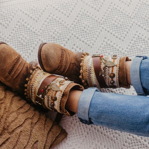 Könnte beinhalten: Braune Wildlederstiefel mit aufwendigen Perlen- und Lederelementen. Die Stiefel werden mit hochgekrempelten blauen Jeans getragen.