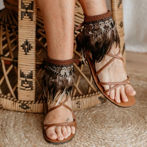 Könnte beinhalten: Braune Ledersandalen mit Knöchelriemen und Federschmuck. Die Sandalen haben ein gewebtes Design und sind mit Metallperlen und Münzen verziert.