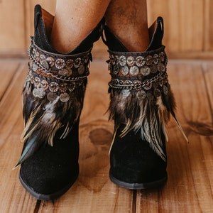 May include: A pair of black ankle boots featuring decorative cuffs adorned with metallic coin-like charms, intricate chain links, and layered brown and black feathers. The boots have a pointed toe and a dark suede-like finish on the exterior.