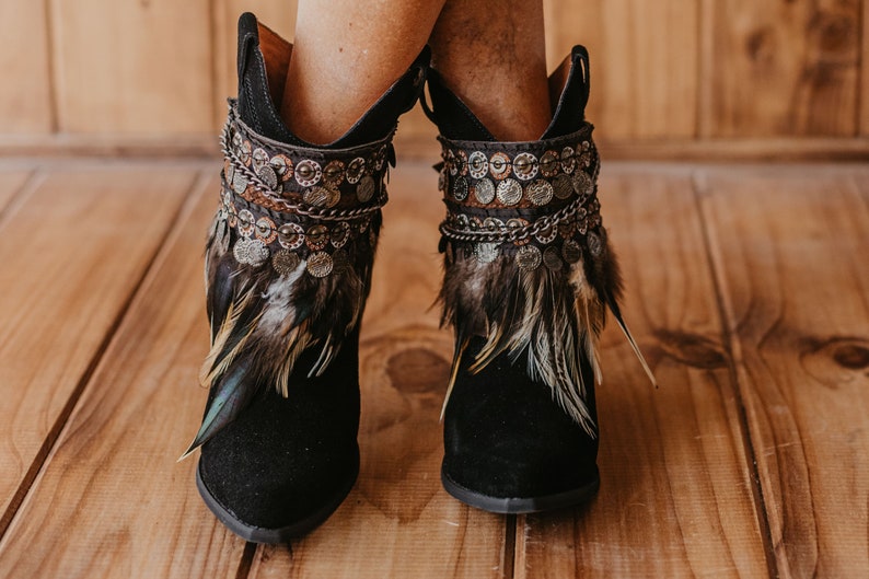 May include: A pair of black ankle boots featuring decorative cuffs adorned with metallic coin-like charms, intricate chain links, and layered brown and black feathers. The boots have a pointed toe and a dark suede-like finish on the exterior.