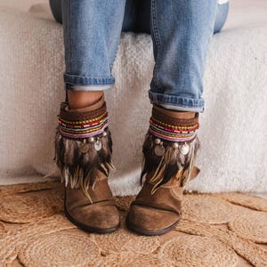May include: Brown suede ankle boots with colourful beaded and feathered ankle cuffs. The boots are being worn by a person wearing blue jeans.