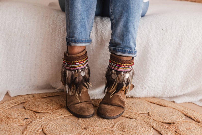 May include: Brown suede ankle boots with colourful beaded and feathered ankle cuffs. The boots are being worn by a person wearing blue jeans.