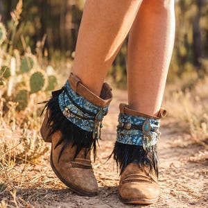 Könnte beinhalten: Braune Wildleder-Stiefeletten mit einer blauen gemusterten Stoffmanschette, schwarzem Federbesatz und hängenden Metallanhängern mit türkisfarbenen Akzenten. Die Stiefel haben einen Blockabsatz und ein geflochtenes Detail am Vorderfuß.