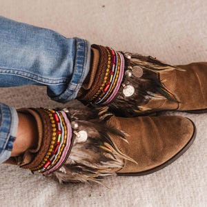 May include: Brown suede ankle boots with feather and beaded embellishments. The boots have a layered look with multiple beaded bracelets and a fringe of feathers.