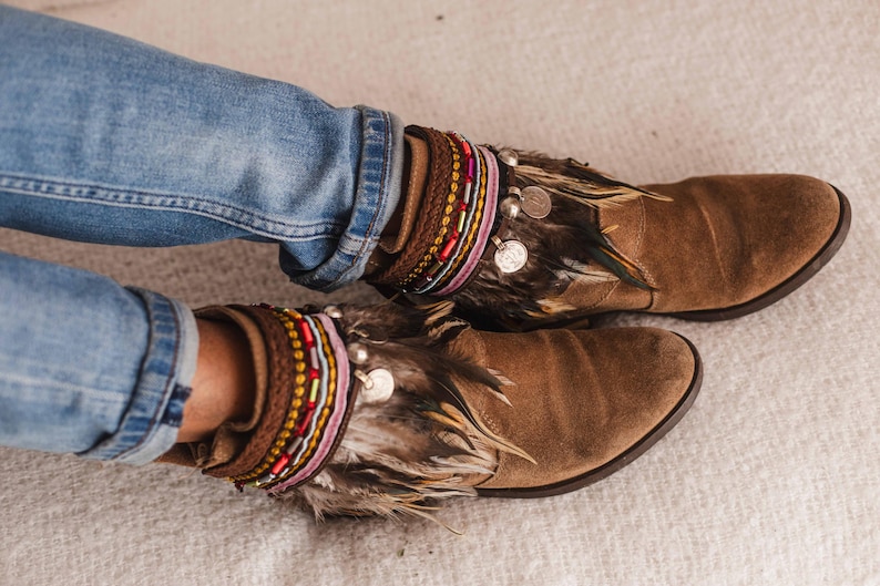 May include: Brown suede ankle boots with feather and beaded embellishments. The boots have a layered look with multiple beaded bracelets and a fringe of feathers.